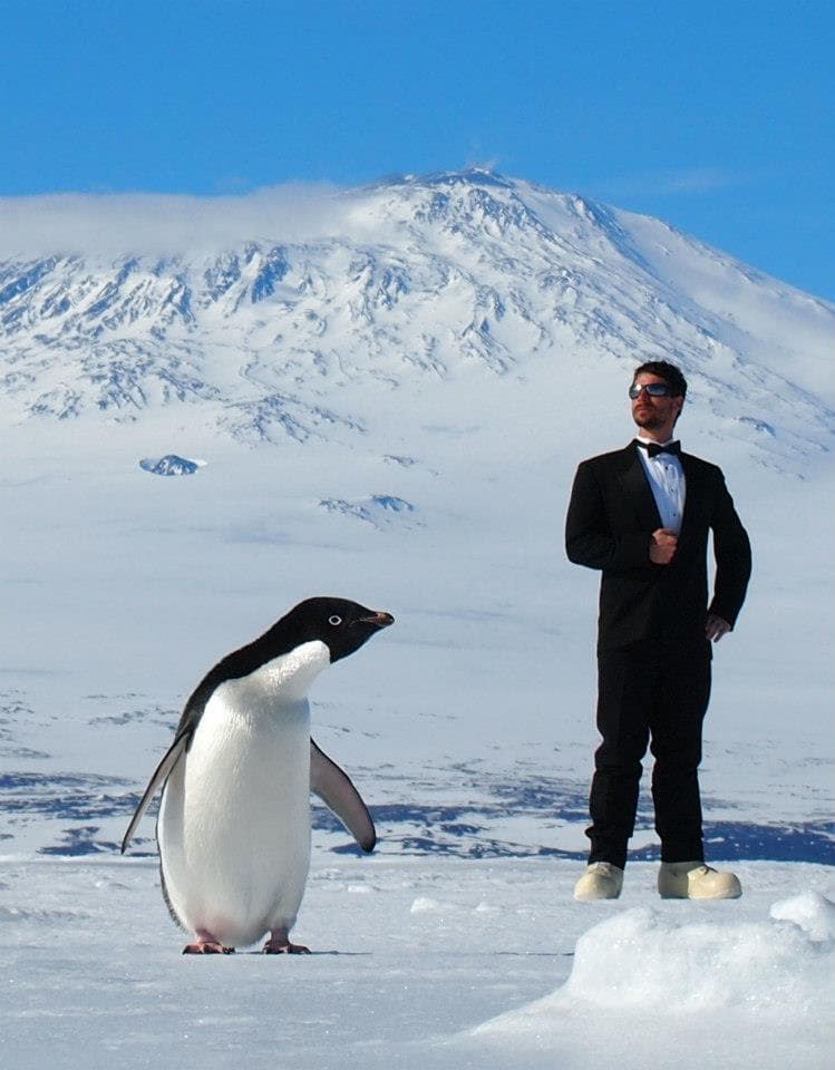 Eric Stackpole in formal attire and cold-weather boots on Antarctic ice next to an Adélie penguin, with a distant snow-covered mountain.