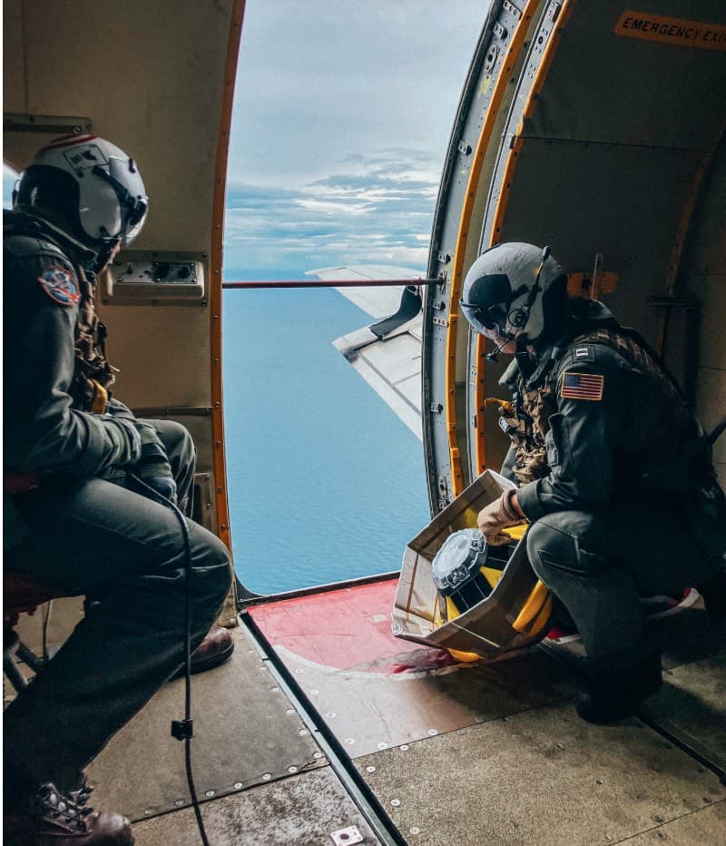Two crew members in flight suits and helmets at an aircraft's open door over the ocean, one holding a yellow Sofar Spotter buoy in a box for deployment.