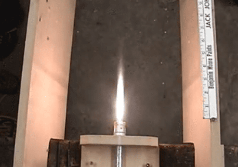 Top-down view of a hybrid rocket motor test firing, with a bright vertical flame framed by two light vertical panels and a ruler along the edge on a dark surface.