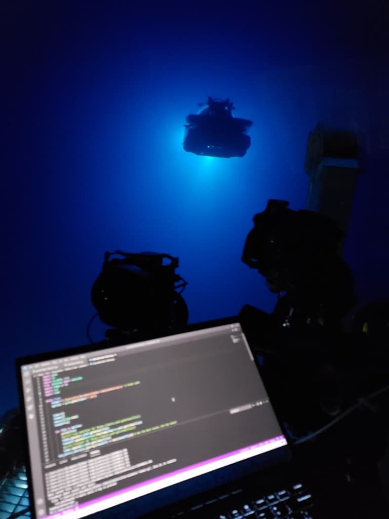 Laptop showing code in the foreground with a lit submersible suspended in deep blue water in the background.