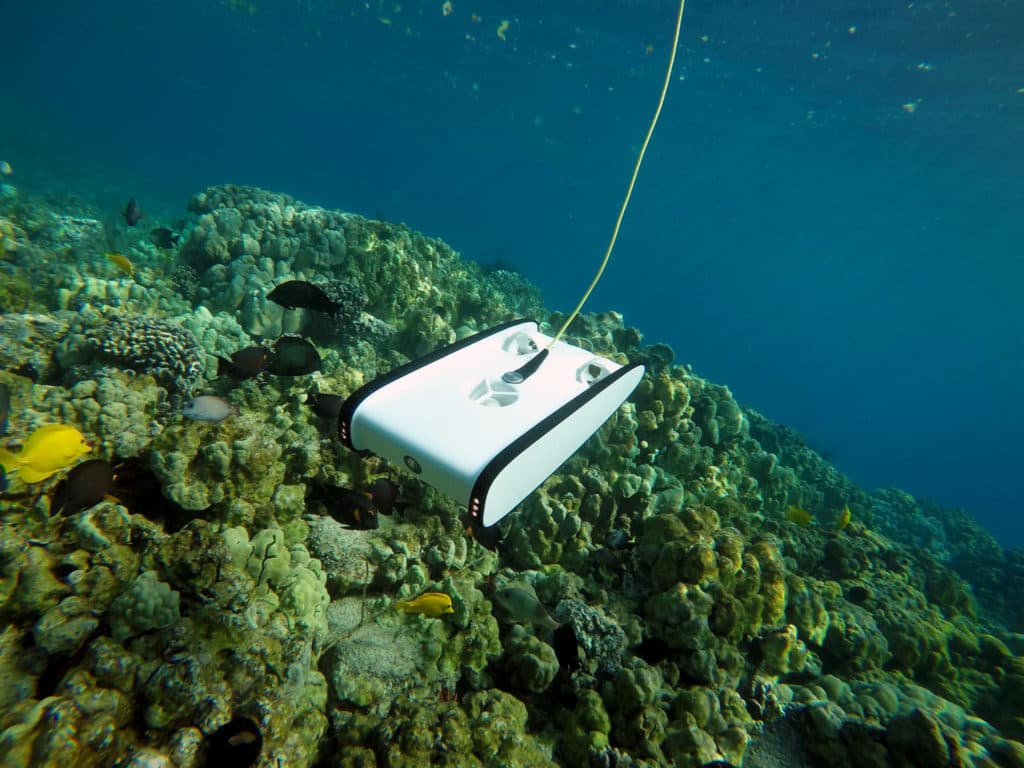 Underwater ROV with yellow tether hovering over a coral reef and tropical fish in clear blue water.
