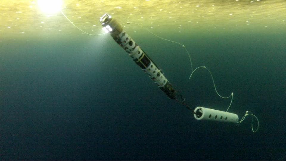 A long cylindrical under-ice instrument illuminated beneath an ice ceiling in dark blue water.
